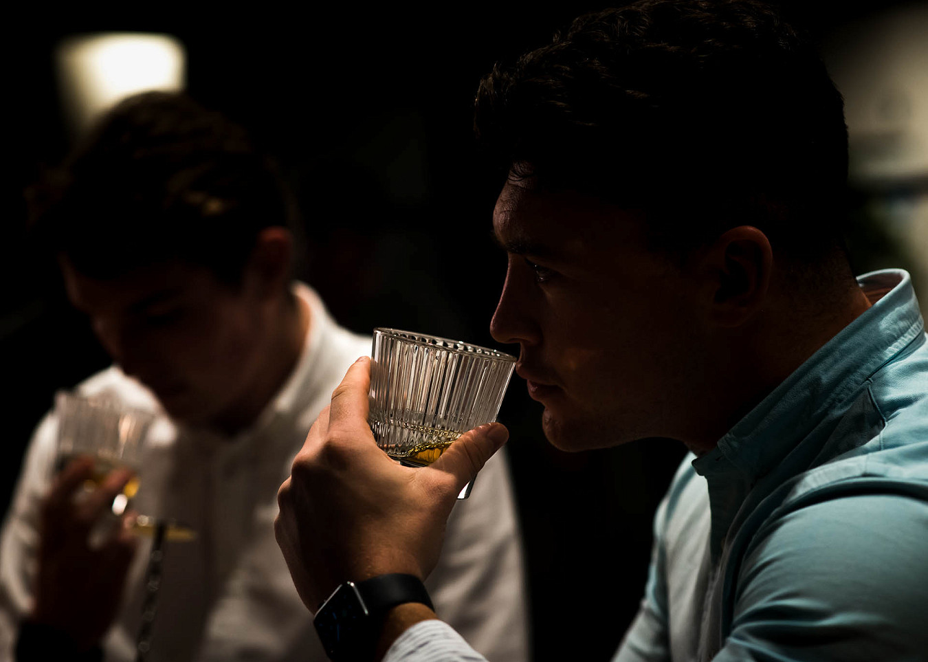Atmospherically lit photo of two men holding glasses of whisky to their noses to smell before drinking