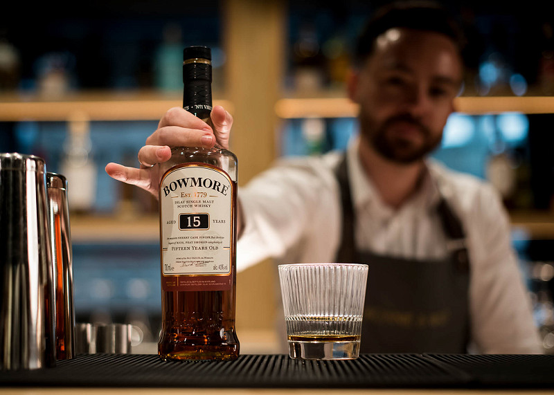 Bartender holding and placing a bottle of Bowmore whisky on a bar with a glass of whisky next to it
